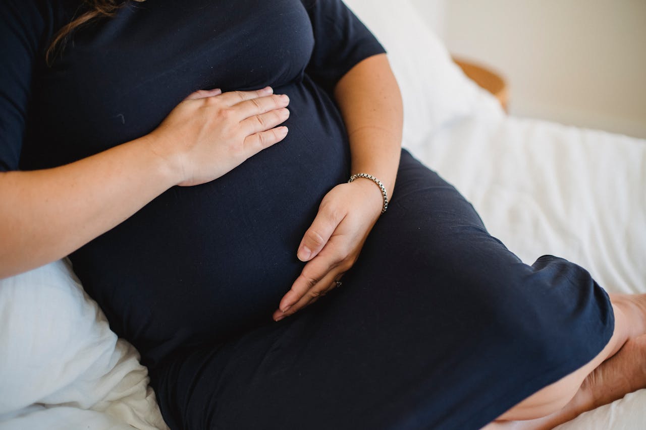 High angle of crop anonymous expectant female in dress stroking tummy while resting on soft bed in house