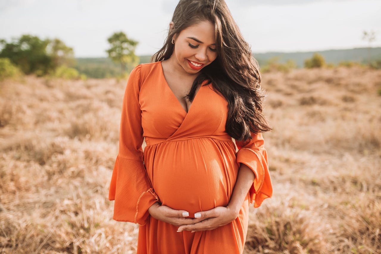 Smiling pregnant woman in an orange dress standing in a serene field during the day.